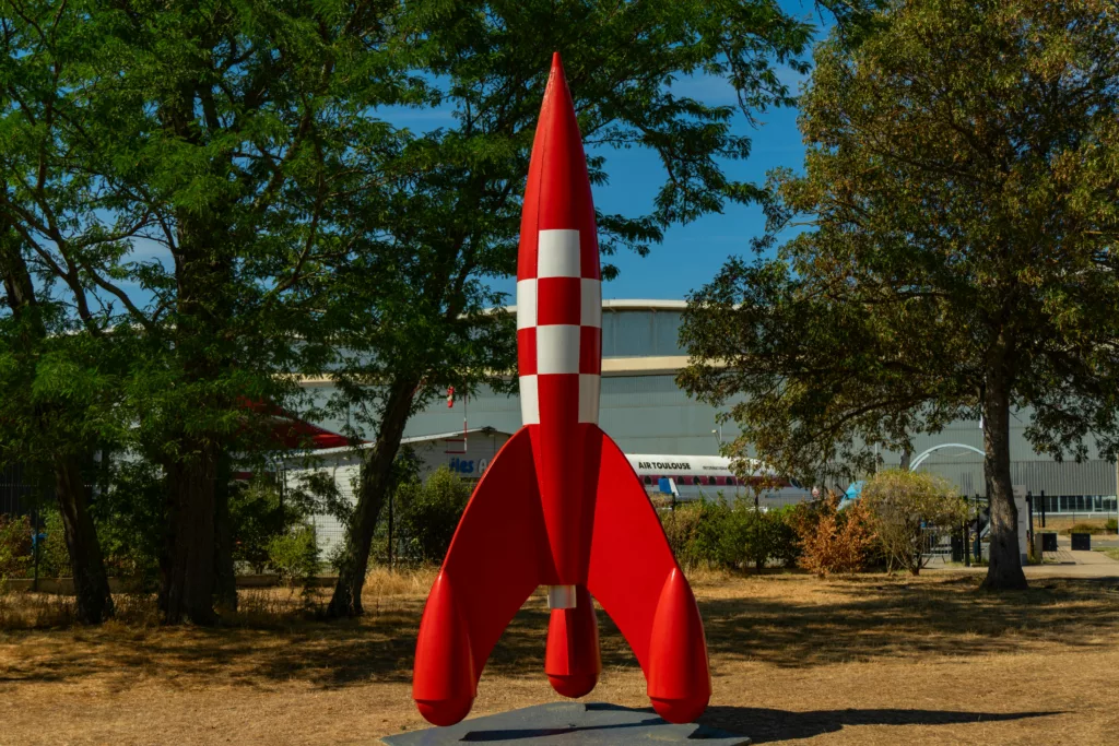 A striking red model rocket displayed outdoors in a sunny park setting in Toulouse, France.